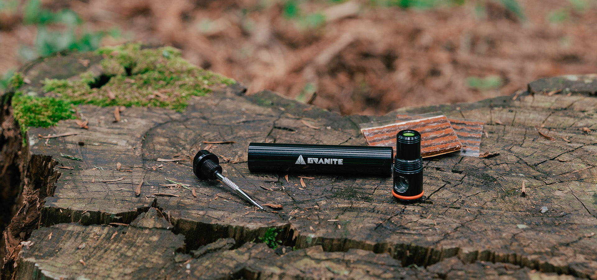 A bike tool kit with a black cylindrical case, multiple repair tools, and a patch kit lying on a mossy tree stump outdoors.