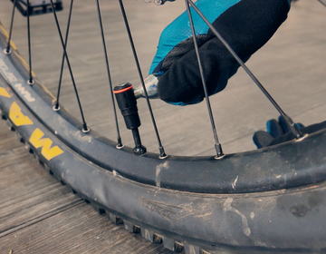 A person wearing a blue and black glove uses a tool to check the air pressure or inflate a mountain bike tire near Granite rocks, focusing on the wheel spokes and valve.