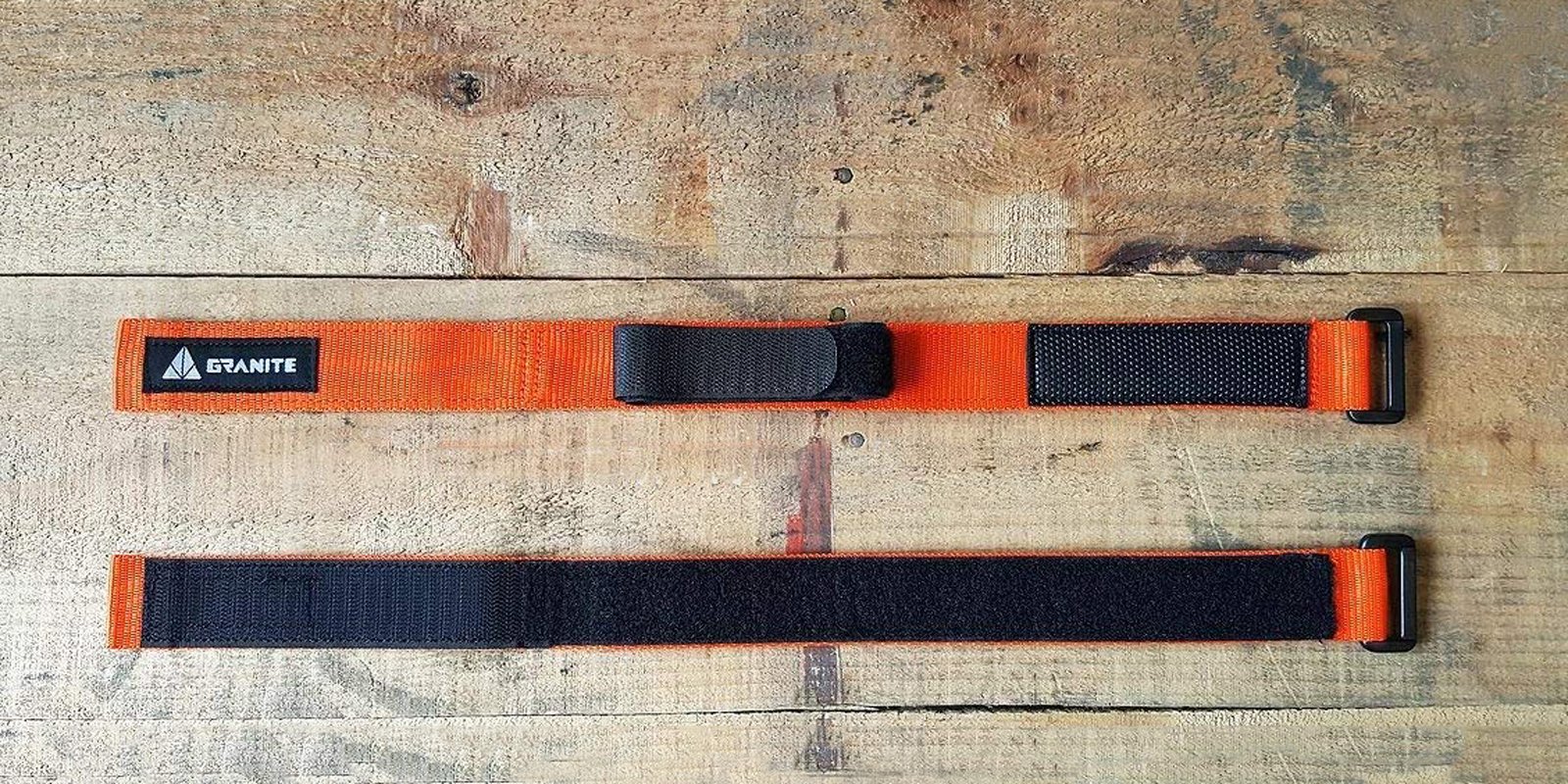 Two orange nylon straps with black Velcro fasteners are laid out on a wooden surface. One strap displays a black Granite brand label, and both have black plastic loops at one end.