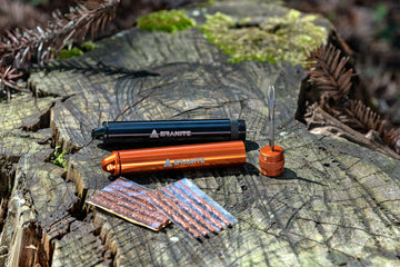 Two compact Granite bike tire repair kits, one black and one orange, sit on a mossy tree stump with several brown tire plugs and a metal insertion tool displayed beside them. Pine needles and forest debris surround the stump.