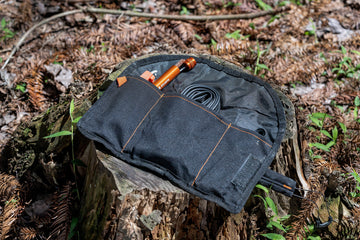 A black fabric pouch with several compartments, holding an orange flashlight, a small canister, and a coiled black cable, sits open on a granite-topped tree stump in a forest setting.
