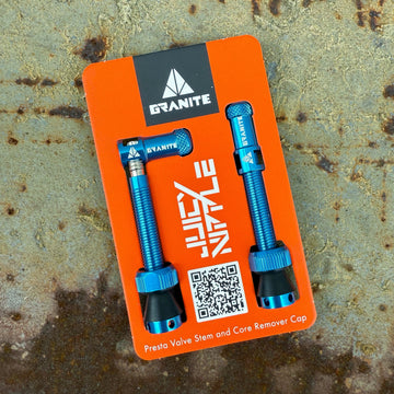 A package of two blue Granite Presta valve stems with core remover caps, shown on an orange backing card with white text and a QR code, resting on a rough, textured surface.
