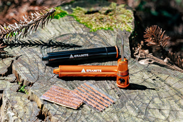 Two Granite tubeless tire repair kits, one black and one orange, rest on a tree stump with tire plugs laid out nearby. Pine needles and branches are scattered around.