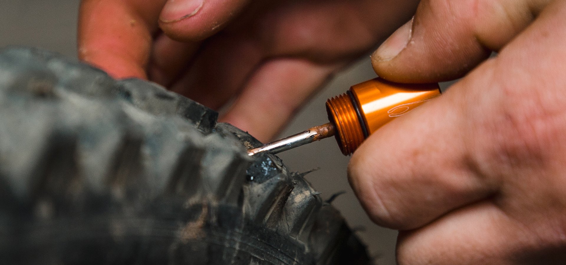 A close-up of hands using an orange tool to apply sealant to a puncture in the worn granite-patterned bicycle tire tread.