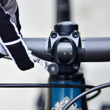 Close-up of a gloved hand using a Granite rocknroll mini ratchet tool on a bike handlebar, tightening the stem bolts securely.