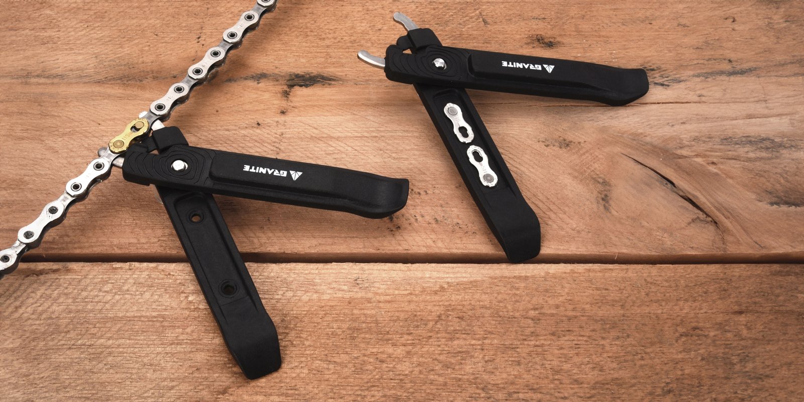 Two black bike chain tools with multi-tool features are placed on a wooden surface next to a partially visible silver bike chain. The tools are open, showing metal components and branding that reads Granite.