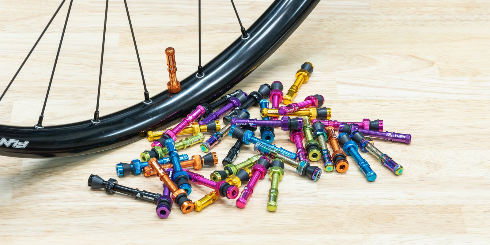 A black bicycle wheel with a copper valve stem sits beside a pile of colorful Granite valve stems scattered on a light wooden floor.