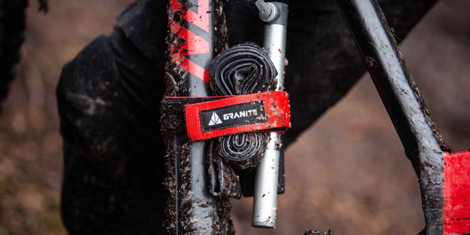 Granite Rockband frame strap in red, securely holding a tube and tool on a muddy bike frame for off-road adventures.