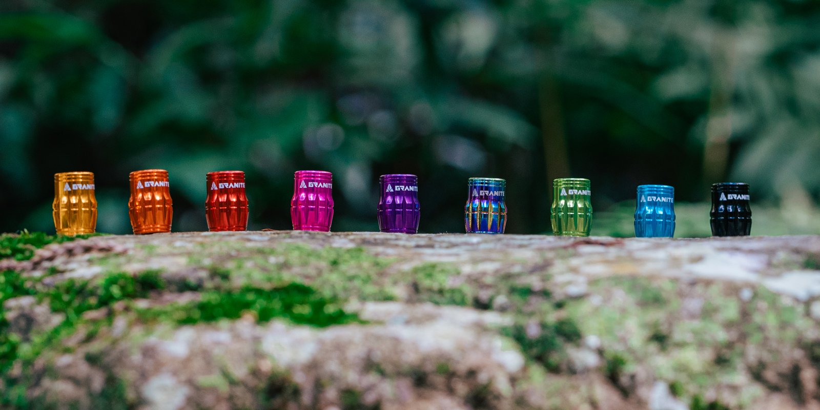 A lineup of colorful Granite Juicy Nipple Schrader valve caps displayed on a mossy log with a blurred green forest background.