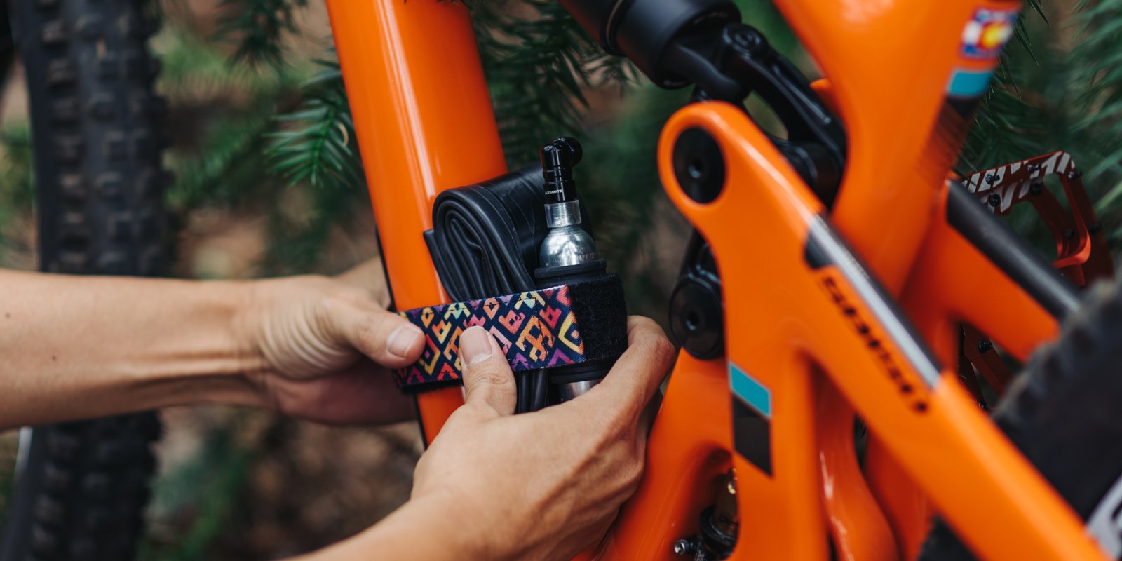 Person securing a spare tube and Punk CO2 inflator to an orange bike frame with a rockband plus strap with square tile pattern.
