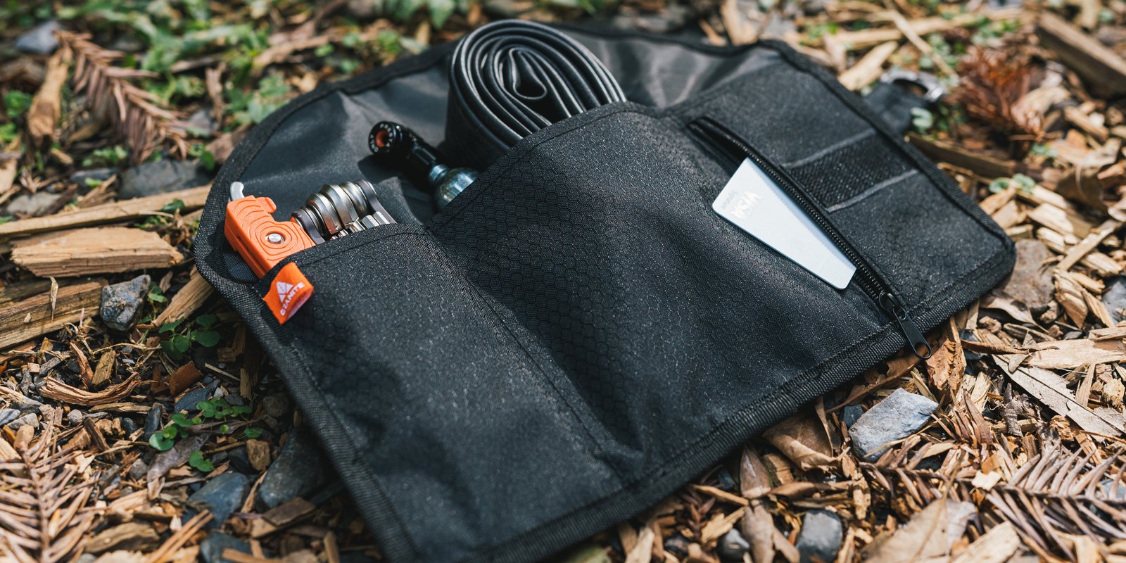 A black tool organizer pouch lies open on the ground among granite stones and wood chips, displaying various tools, a bike inner tube, a folded card, and other small items in its compartments.