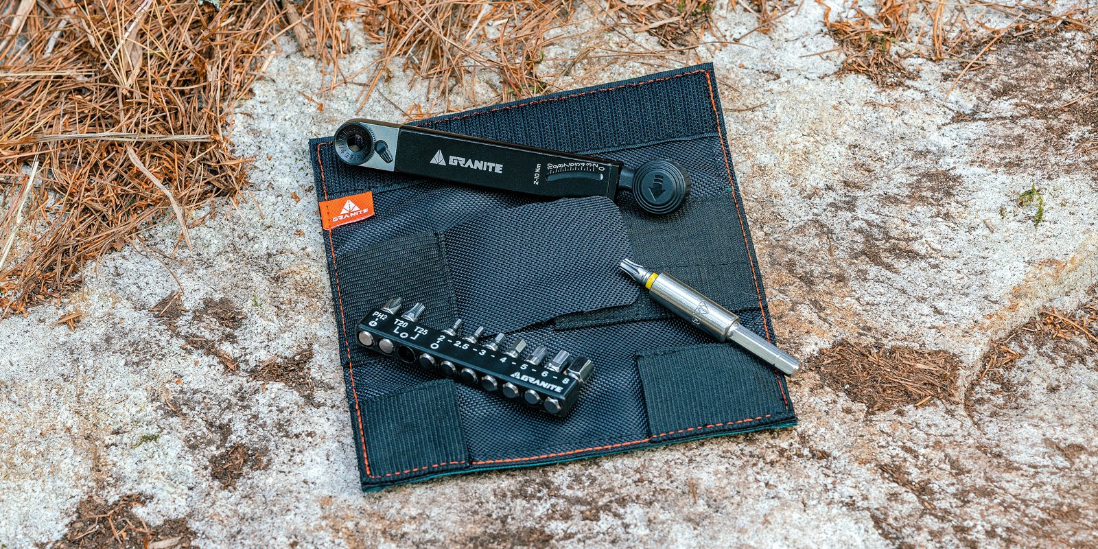A compact Granite bike tool kit, featuring a ratchet wrench, multi-tool with bits, and a small storage pouch, is laid out on a rock surface with pine needles nearby.
