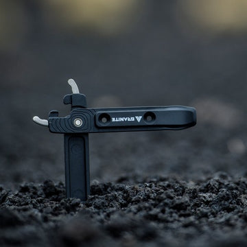 A black Granite-branded multi-tool is partially embedded upright in dark soil, with two metal levers extended. The blurred background draws focus to the tool.
