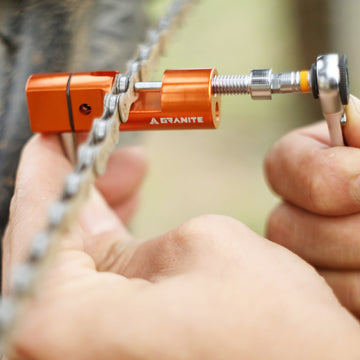Hands using an orange Granite Stash chain tool on a bicycle chain outdoors, driving the rivet out with a small ratchet