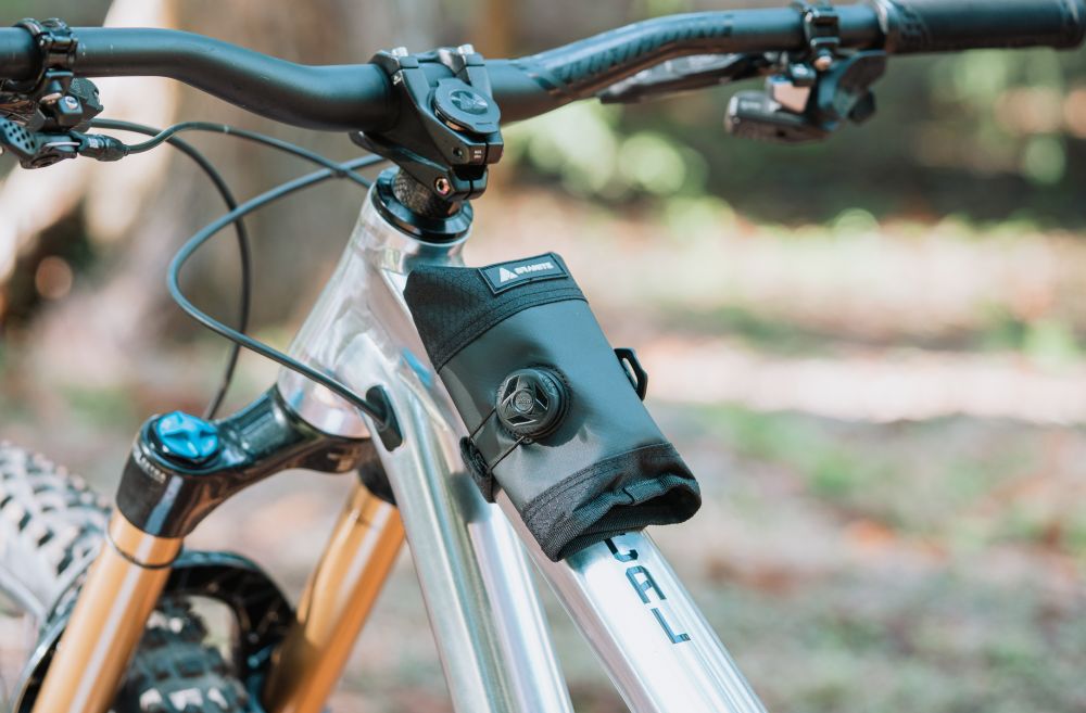 A black bike bag is attached to the top tube and head tube of a silver mountain bike, positioned just below the handlebars in an outdoor setting.