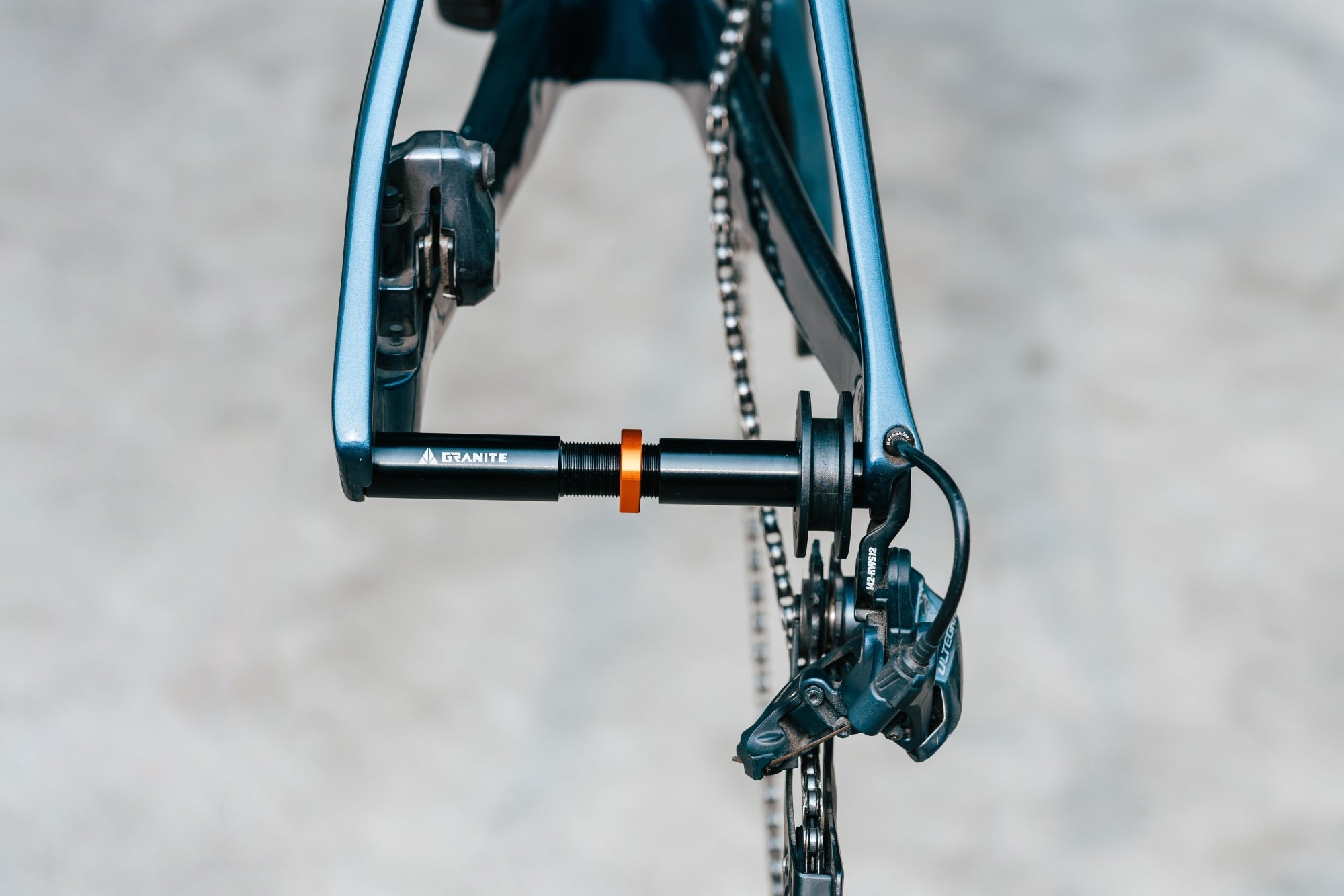 Close-up of a bicycle rear dropout with an axle and derailleur visible, showing part of the chain and frame against a blurred, light-colored background.