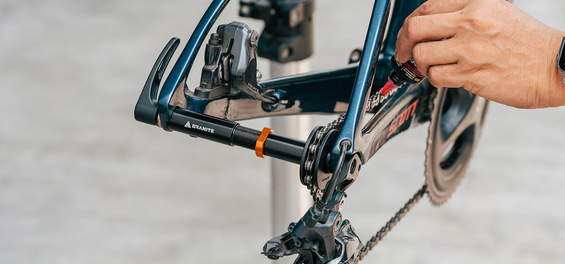 A person lubricates the rear derailleur of a bicycle, which is mounted on a repair stand. The bikes chain and crankset are visible, and the person is holding a small bottle of lubricant.