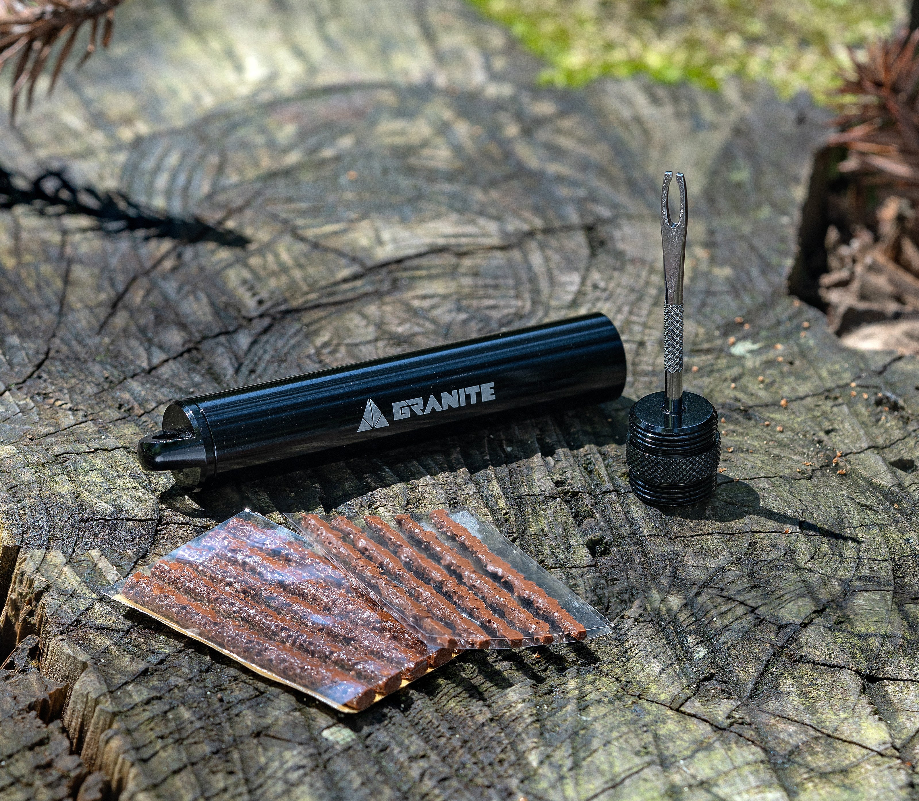 A tire plug kit with a black Granite case, a metal insertion tool, and several brown rubber plugs is displayed on a tree stump outdoors.