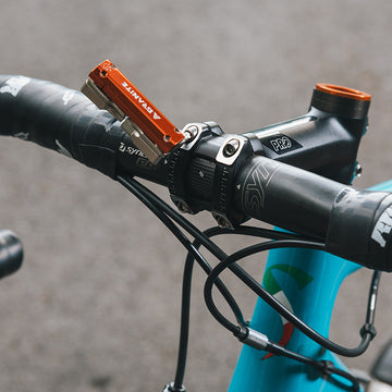 Close-up of a bike handlebar with an orange Granite Stash RCX multi-tool attached, featuring black grips and a sleek blue frame.