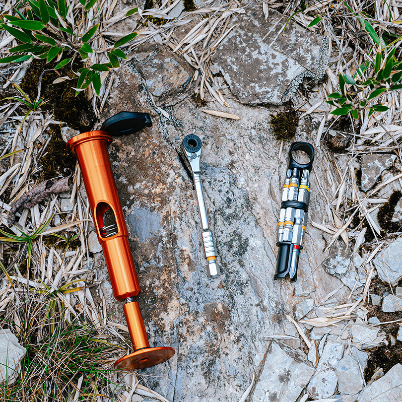 Three bike tools on a rocky surface: an orange Stash RT multi-tool, a silver ratchet, and a black multi-bit tool kit.