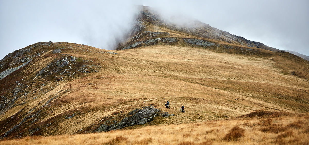 Two cyclists riding across an open grassy mountainside beneath a mist-covered peak
