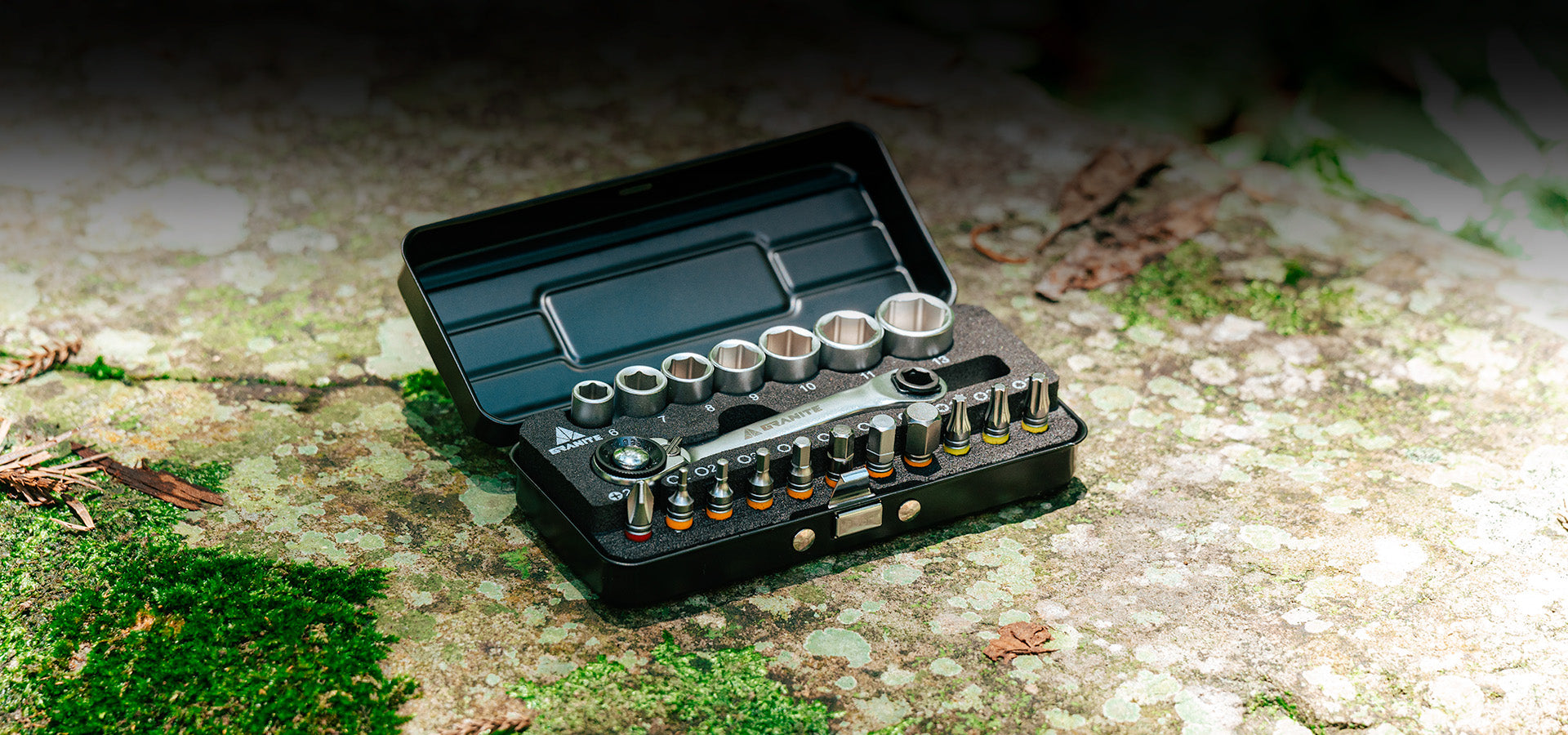 Granite Duet Hex Bit & Socket Ratchet Tool Kit displayed on a moss-covered rock outdoors. The open black case reveals neatly arranged hex sockets, bits, and a ratchet tool, emphasizing its compact and portable design suitable for on-the-go bike repairs in rugged environments.