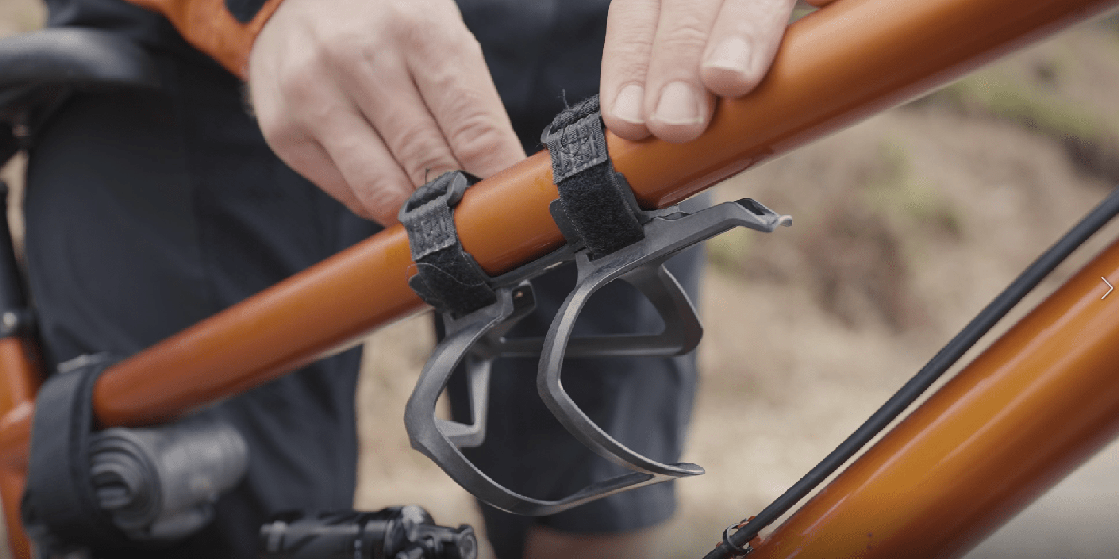 Hands securing Granite AUX Bottle Cage onto an orange bike frame using straps for easy attachment and stability.