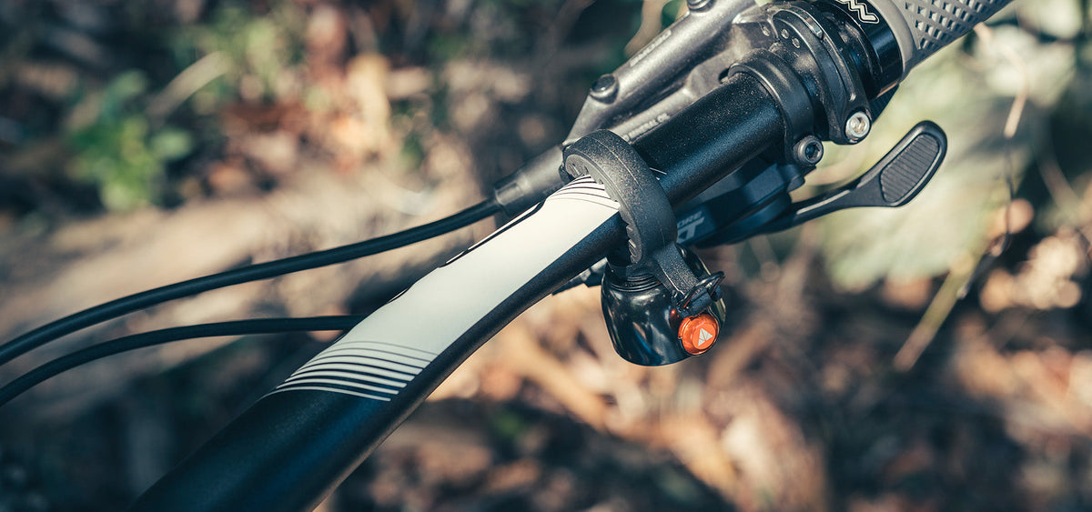 Granite Cricket bike bell mounted near the stem on a mountain bike handlebar with a Shimano XT brake lever, orange striker facing out, blurred autumn forest behind