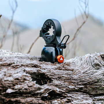 Granite Cricket bicycle bell placed on a rugged tree trunk, featuring a unique mounting design, against a blurred natural background.