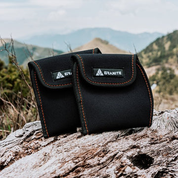 Pair of Granite Pita Pedal Covers with black fabric and orange stitching displayed on a log against a mountainous background.