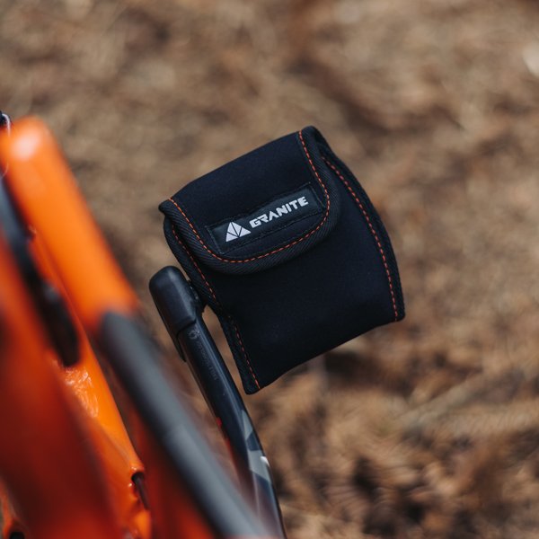 Granite Pita Pedal Cover in black with orange stitching attached to an orange bike frame, protecting the pedal in a rugged outdoor setting.