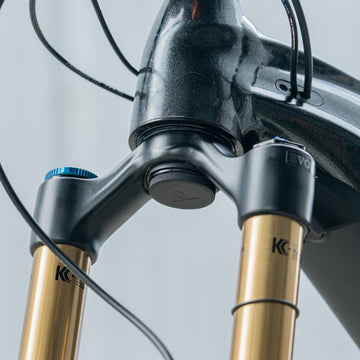 Close-up of a mountain bike’s front suspension fork with gold-colored stanchions and black adjustment knobs, attached to a matte black Granite frame. Several cables are visible around the head tube.