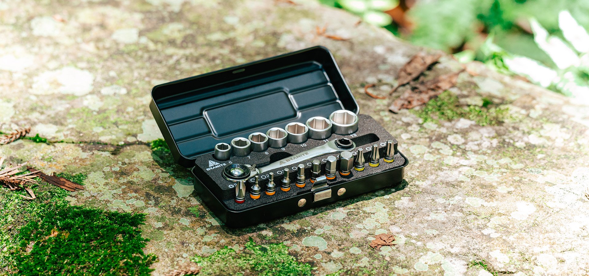 Granite Duet Hex Bit & Socket Ratchet Tool Kit displayed on a moss-covered rock outdoors. The open black case reveals neatly arranged hex sockets, bits, and a ratchet tool, emphasizing its compact and portable design suitable for on-the-go bike repairs in rugged environments.