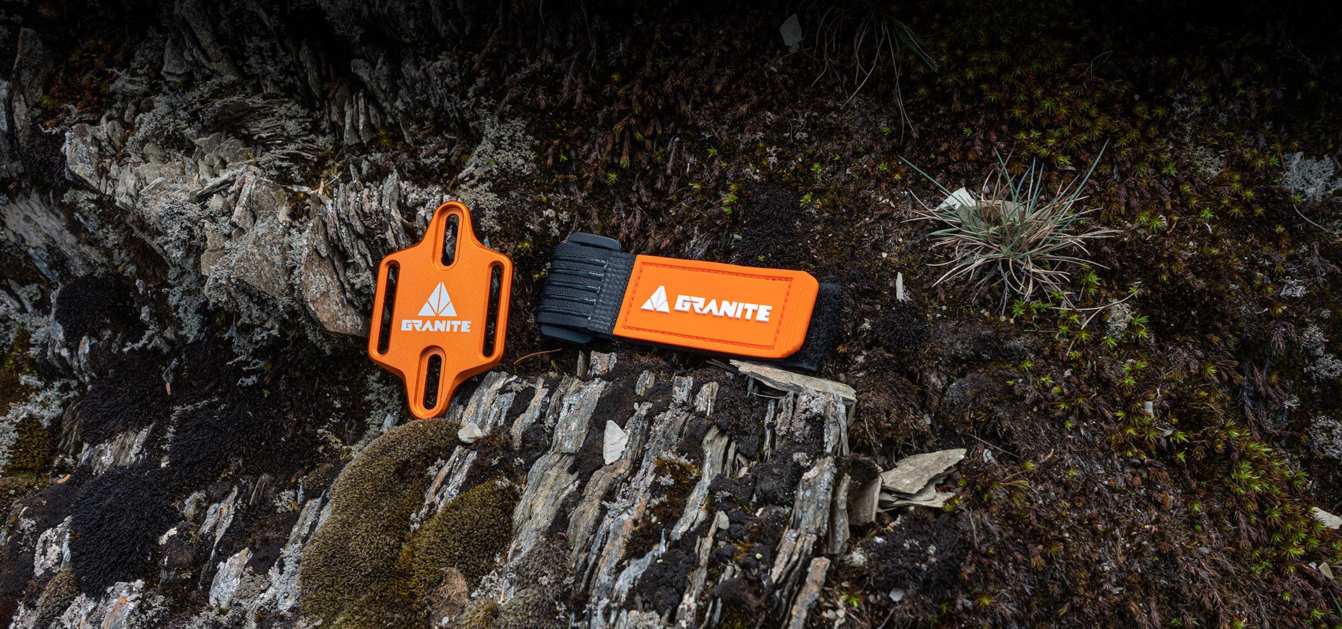 Granite Portaledge bike bottle cage mount strap displayed on a rugged outdoor surface. The orange metal plate and strap, with the Granite logo, are placed on rocky terrain with patches of moss and small plants, emphasizing the product's durability and ruggedness for outdoor use.