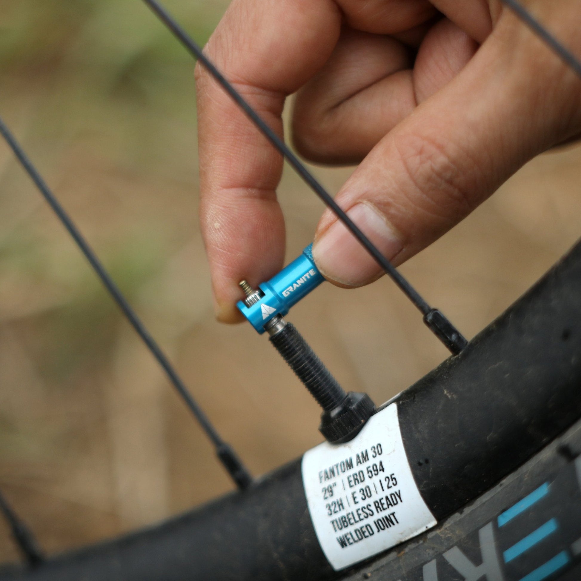 Hand using a blue Granite Presta valve cap tool to remove the valve core from a bike tire on a black rim.