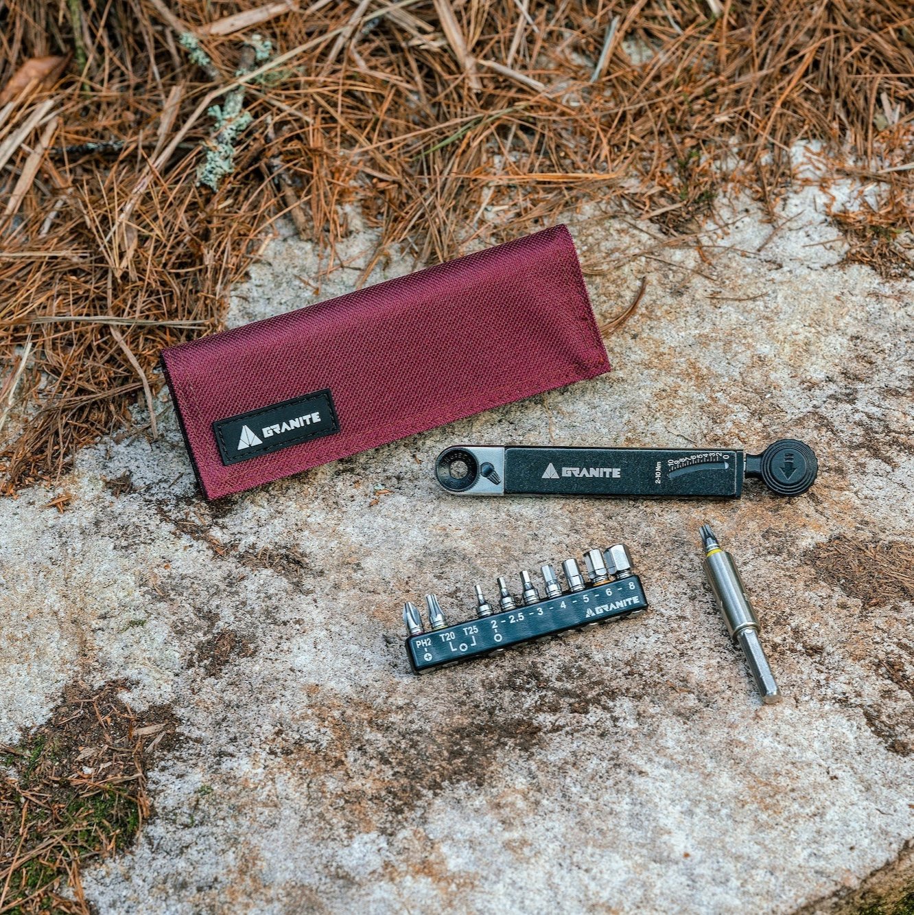 Rocknroll TQ Torque Ratchet Tool with maroon pouch, multitool, bit holder, and bits displayed on a rocky surface outdoors.