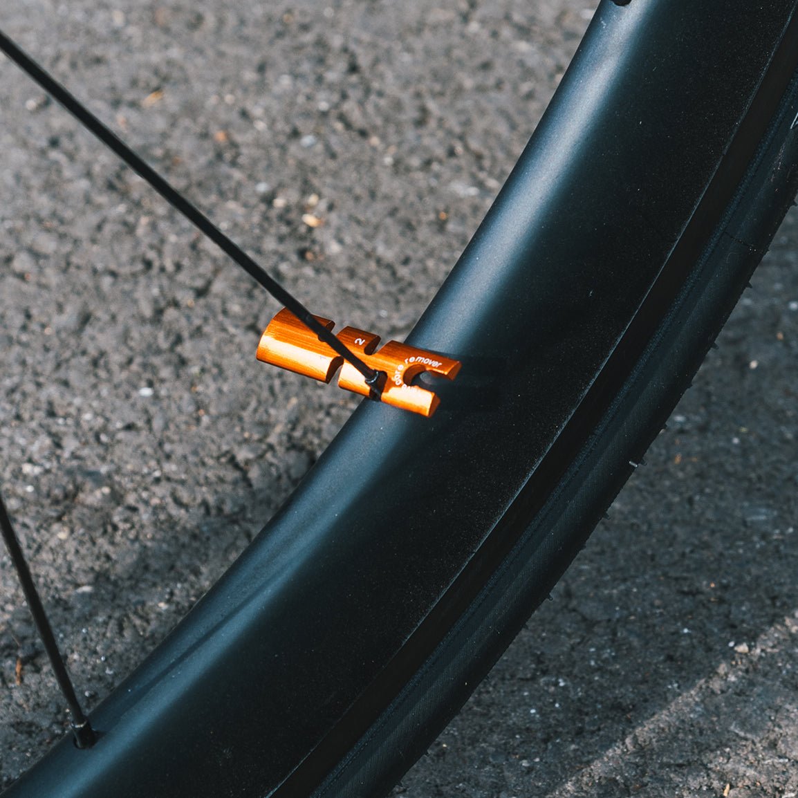 Close-up of an orange Granite spoke tool attached to a bike wheel spoke, with a black rim and asphalt background.