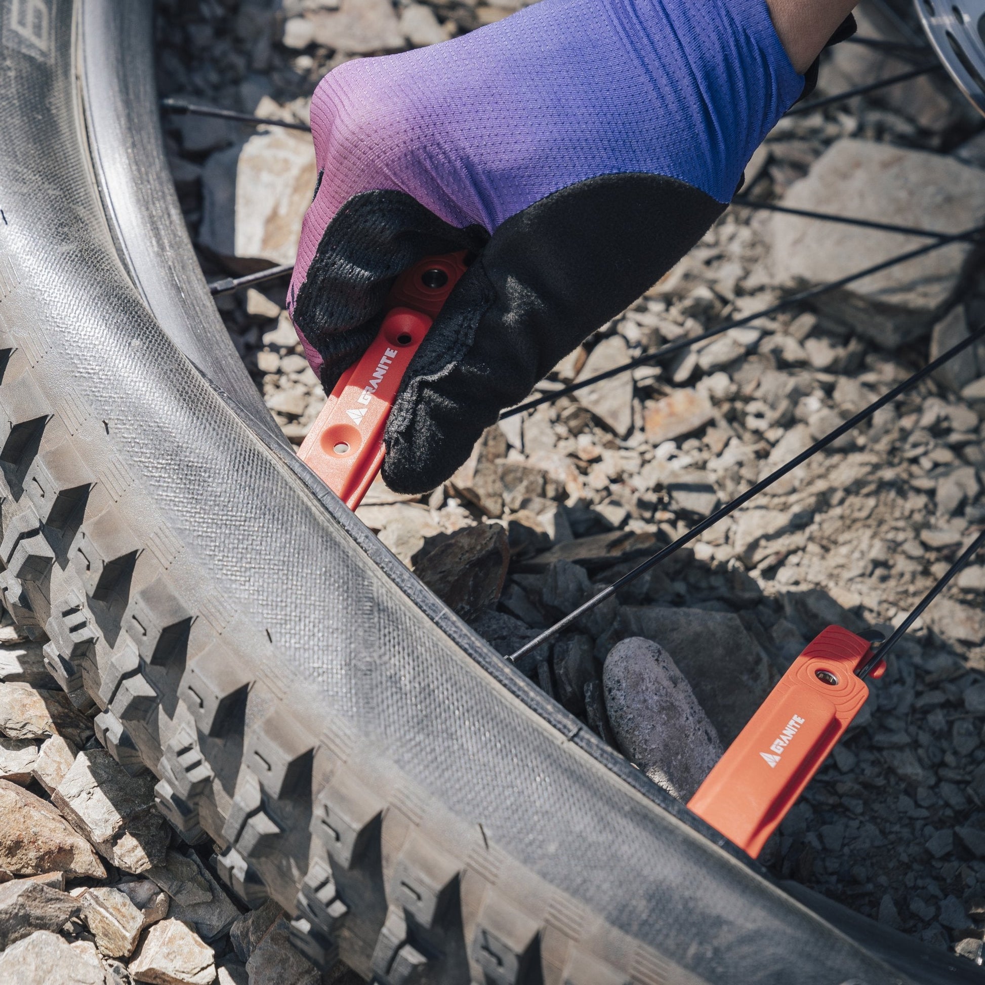 Person using Granite Talon orange tire levers to remove a bike tire on rocky terrain, wearing black and purple gloves.