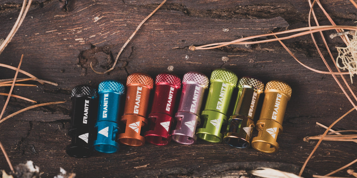 Eight anodised Granite Juicy Nipple Presta valve caps in a row on dark weathered wood: black, blue, orange, red, purple, green, oil slick and gold.