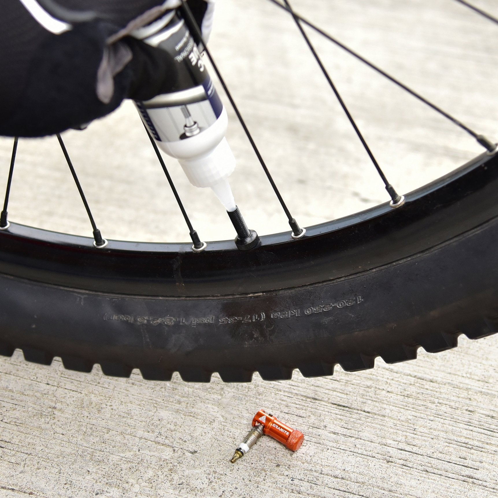 Cyclist applying sealant to a bike tire valve stem with an orange Granite valve cap lying nearby on the ground.