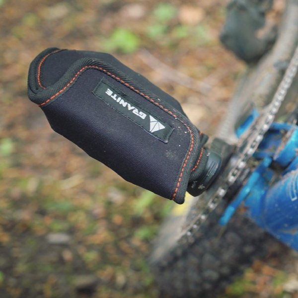Close-up of a black protective cover with orange stitching labeled Granite on a bicycle pedal, with part of a blue bike frame and the ground in the background.