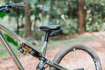 Black Granite Quiver tool roll rolled up under the saddle of a raw silver Commencal full-suspension mountain bike, seen from behind on a forest trail