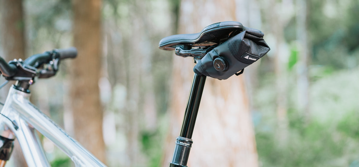Black Granite Quiver tool roll strapped under the saddle of a silver mountain bike on a BikeYoke dropper post, ratchet dial facing out, blurred forest behind