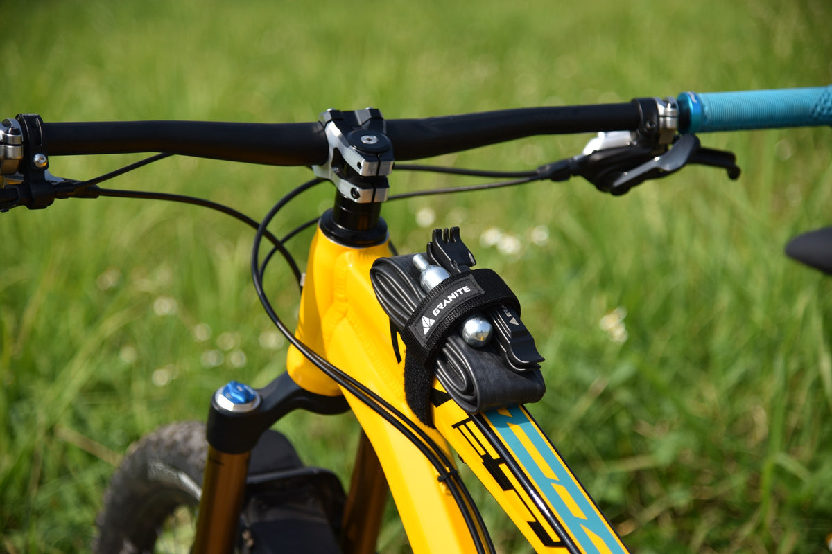 Black Granite Rockband strap holding a spare tube, two CO2 cartridges and a tool on the top tube of a yellow mountain bike, green grass behind