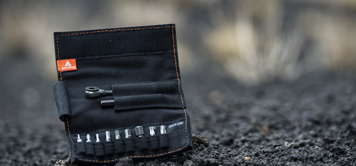 Open black Granite Rocknroll tool roll showing a mini ratchet, extension rod and nine bits, standing on dark volcanic gravel