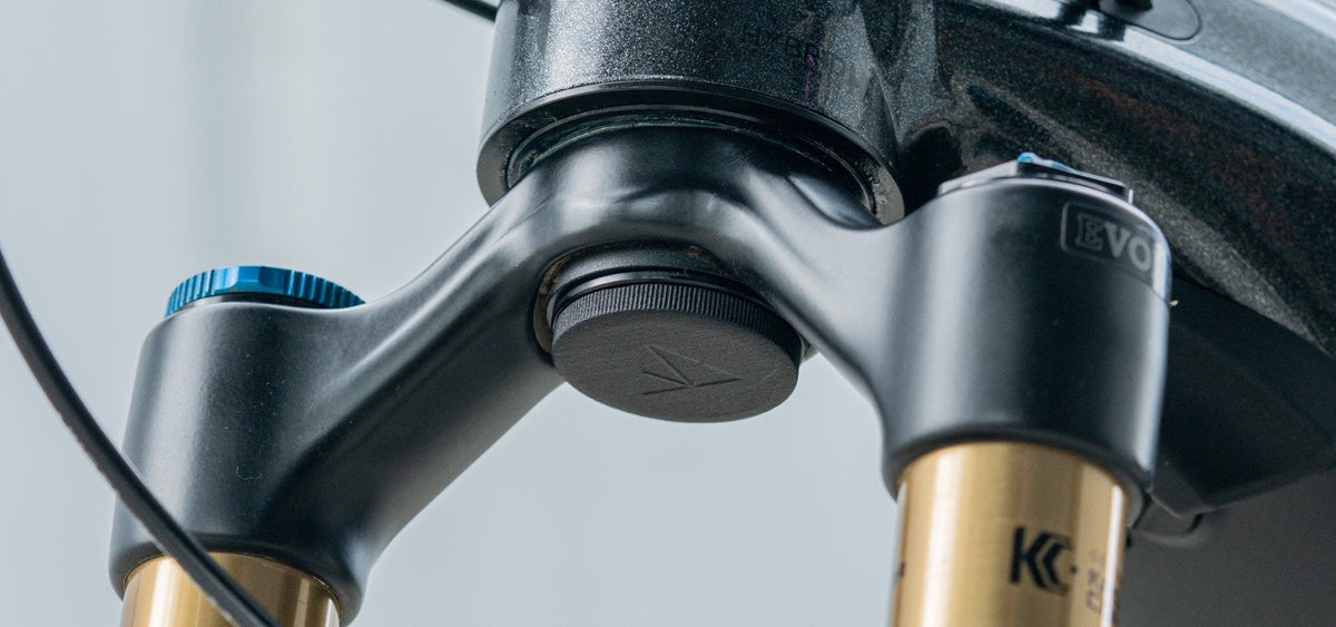 Granite Stash AirTag Mount installed as the black lower cap under a mountain bike fork crown, gold suspension stanchions either side, showing the Granite arrow logo on the cap