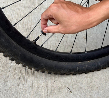 A close-up of a hand unscrewing a valve core from a bicycle tire using a valve core tool. The tire is a large mountain bike tire, and the valve core has been partially removed from the Presta valve, with the removed part lying on the concrete surface below the tire.